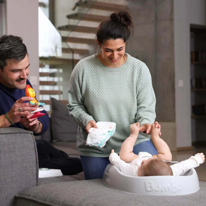 Woman changing a baby's diaper in a living room with a man holding a toy.