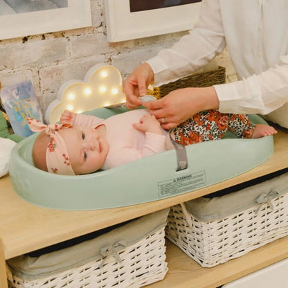 Baby lying in a green bassinet with a person adjusting something above, in a home setting.
