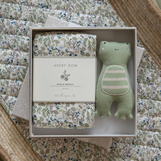 Green plush toy and swaddle blanket in a box with 'Avery Row' branding on a floral fabric background.