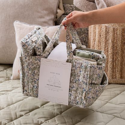 Floral-patterned baby organizer held by a hand on a bed with pillows and blankets.