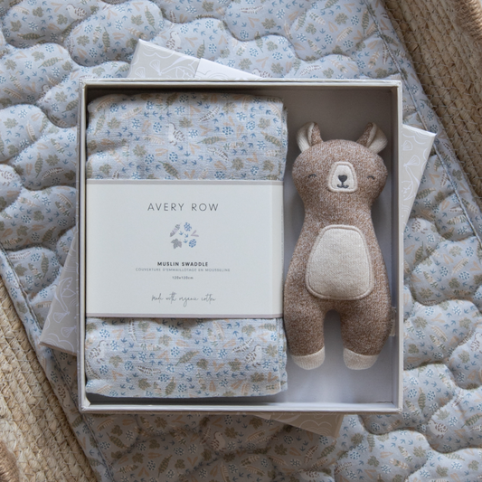 Baby gift set with swaddle blanket, plush toy, and card in a box on a textured surface.