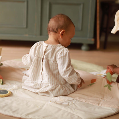Baby in a white outfit sitting on a beige blanket with toys around