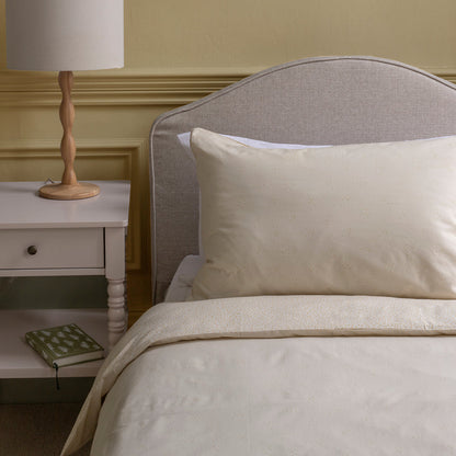 Neatly made bed with beige pillows and duvet, nightstand with lamp and book in a bedroom setting.