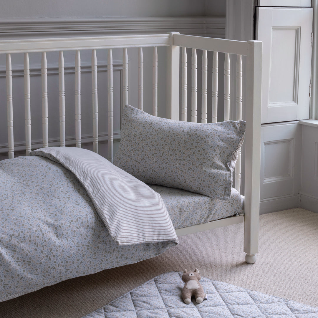 White crib with gray bedding and pillow in a room with white walls and carpet.
