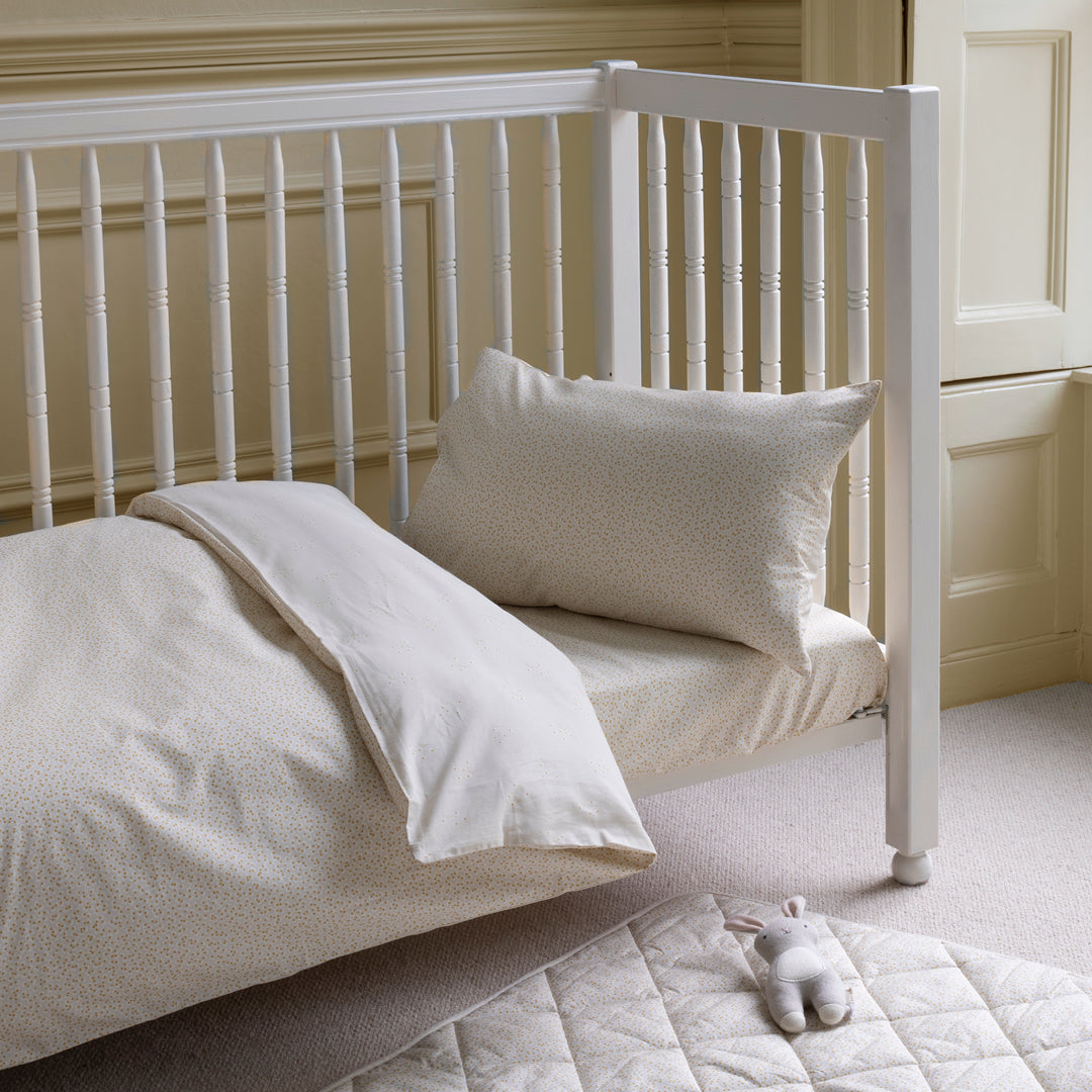 White crib with beige bedding and a pillow in a room with light-colored walls and flooring.