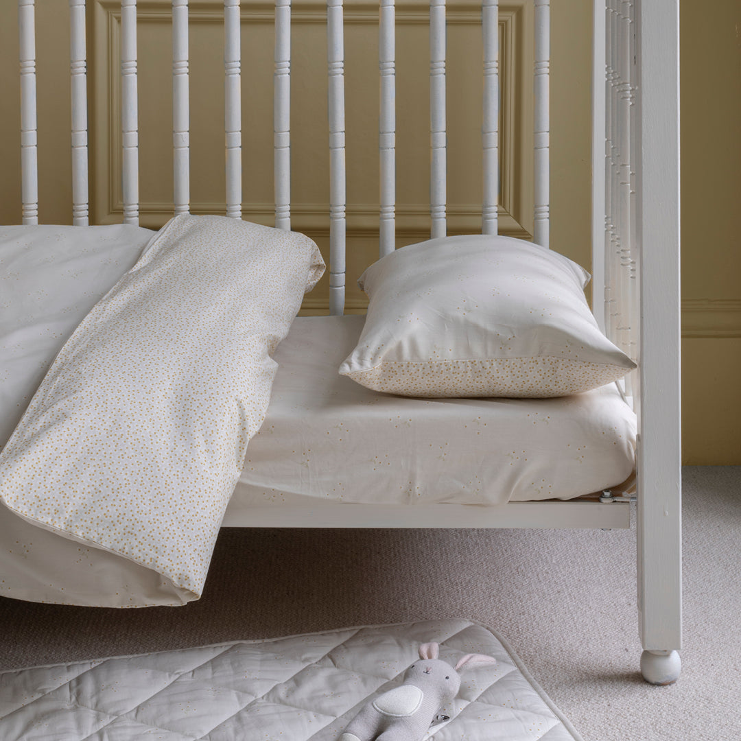 White crib with bedding and a toy on a carpeted floor against a beige wall.