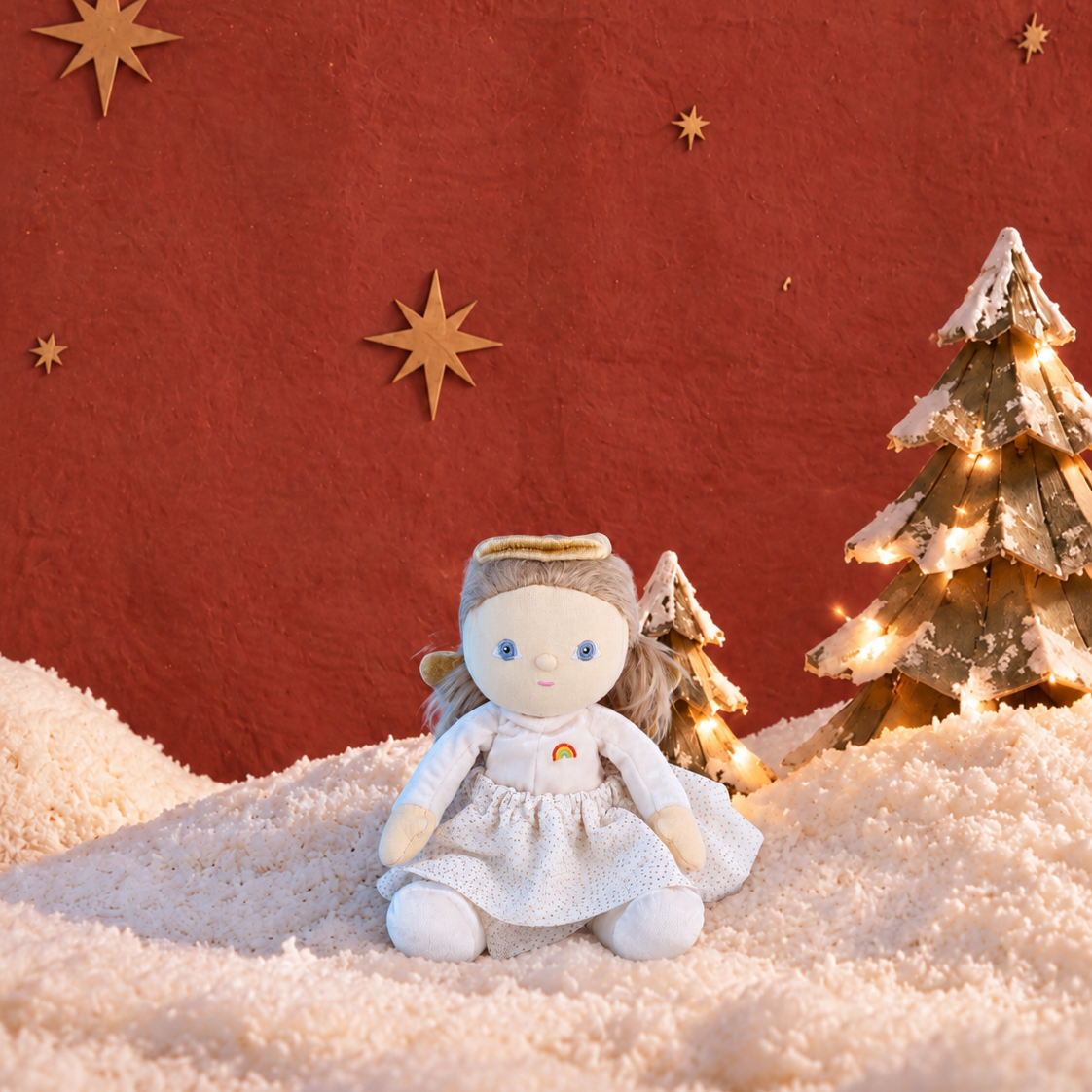 A small white angel doll with a gold halo sits on a snowy surface against a textured red-orange background, with decorative gold stars and two small lit Christmas trees.