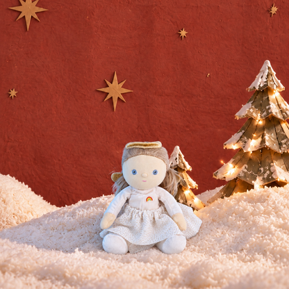 A small white angel doll with a gold halo sits on a snowy surface against a textured red-orange background, with decorative gold stars and two small lit Christmas trees.