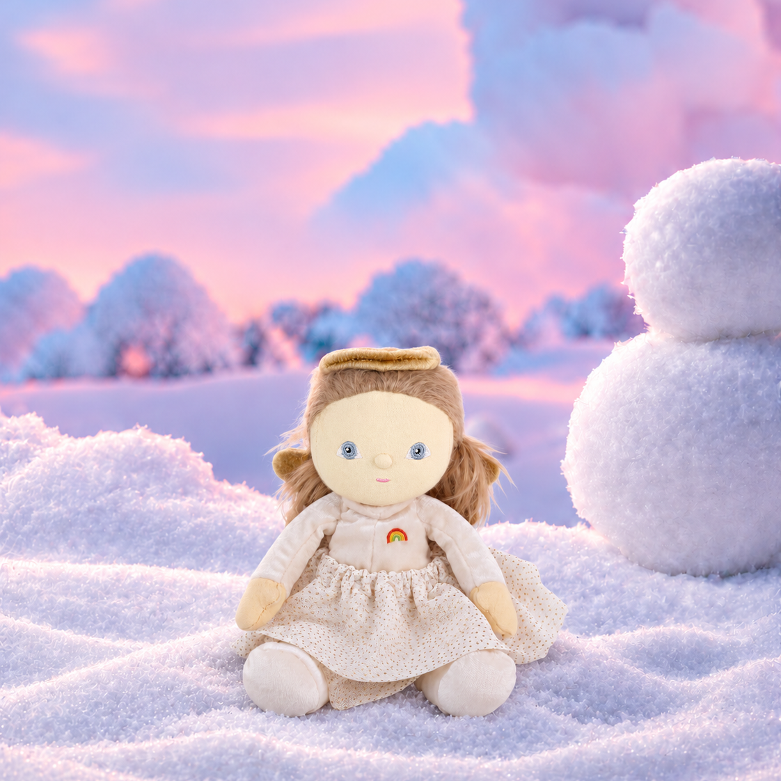 A small plush doll with light brown hair sits in a snowy outdoor scene with a pink and blue sky, trees, and a snowman to the right.