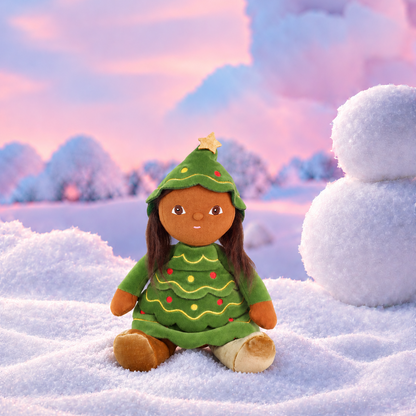 A plush doll with brown skin and long dark hair sits in the snow wearing a green Christmas tree dress with yellow and red ornaments and a gold star on the hat. A snowman stands to the right, with a soft pink and blue winter sky and trees in the background.