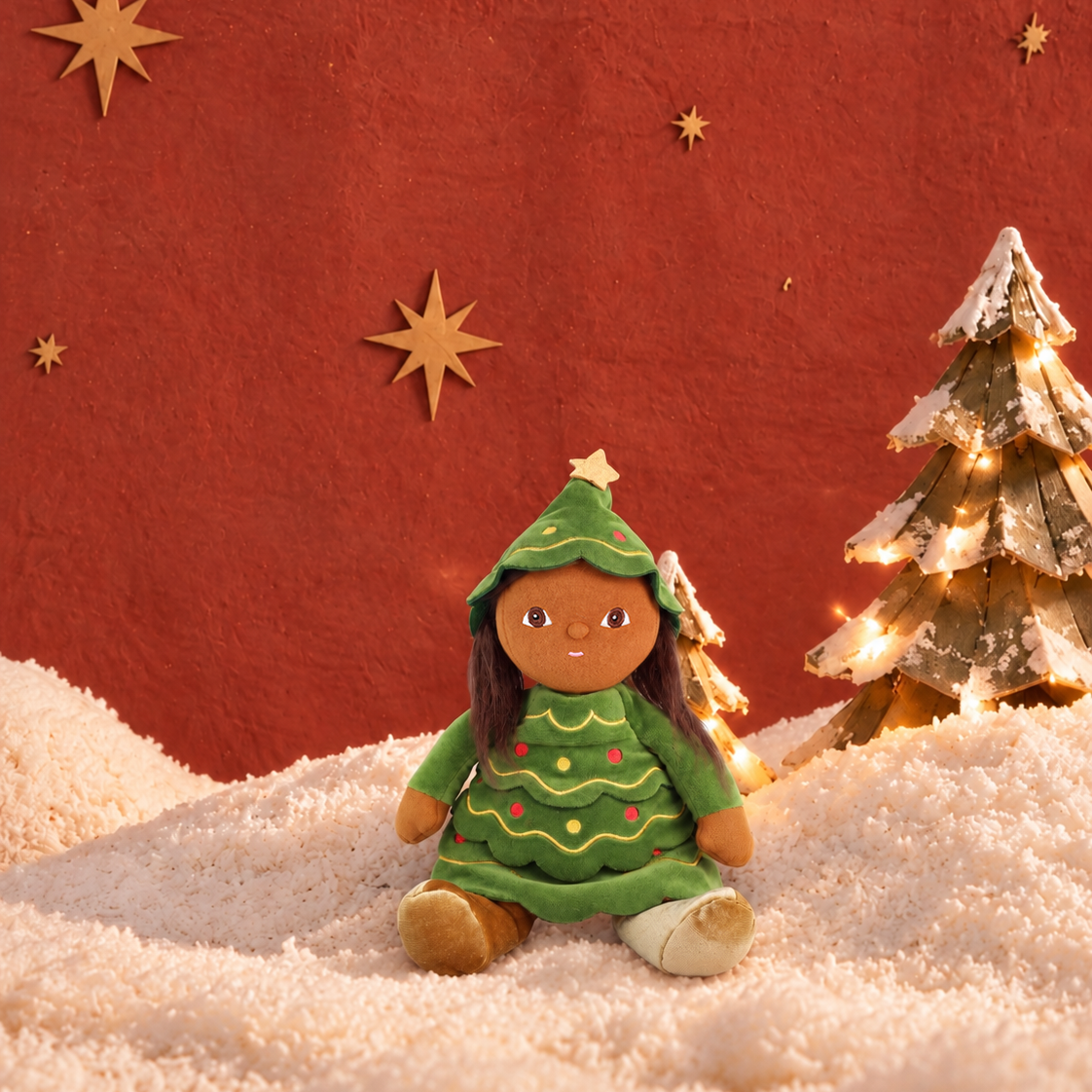 A small doll dressed in a green Christmas tree outfit sits on a textured white snowy surface against a warm red wall, with star decorations and a lit gold Christmas tree in the background.