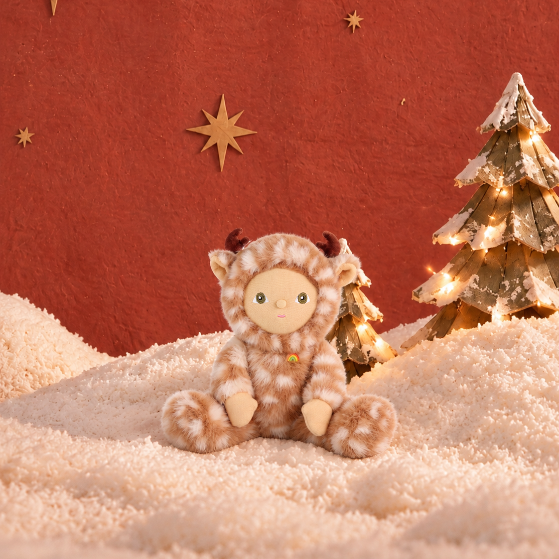 A plush toy dressed as a reindeer sits on a soft cream faux-fur surface against a textured rust-red backdrop, with small star cutouts and a decorated Christmas tree to the right.