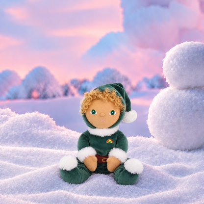 A small plush elf doll with curly blond hair sits in the snow wearing a dark green outfit with white trim, set against a dreamy winter landscape with a snowman and pastel sky.