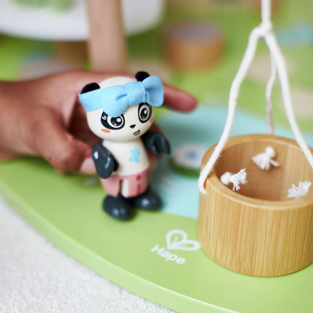 Child's hand holding a panda toy with a blue bow, next to a wooden toy with hanging elements on a green surface.