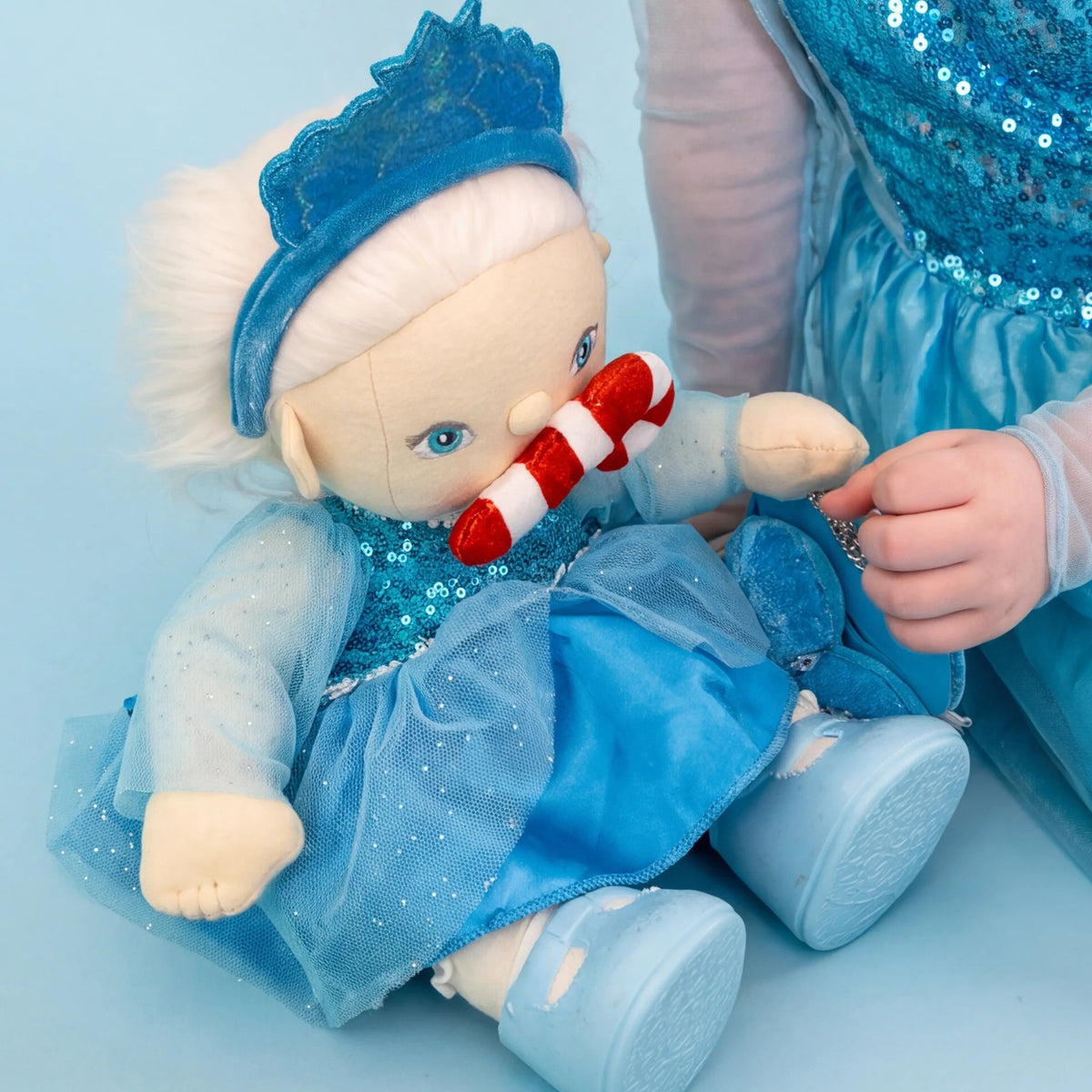 A plush doll dressed in a blue sequined gown and matching blue crown, seated on a light blue background. A child wearing a matching blue sequined dress is partially visible on the right, holding the doll.