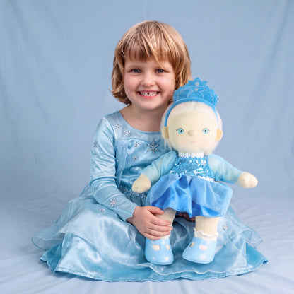 A young child with blonde hair sits on a light blue fabric backdrop, wearing a shiny light blue dress and holding a matching blue doll with a crown.