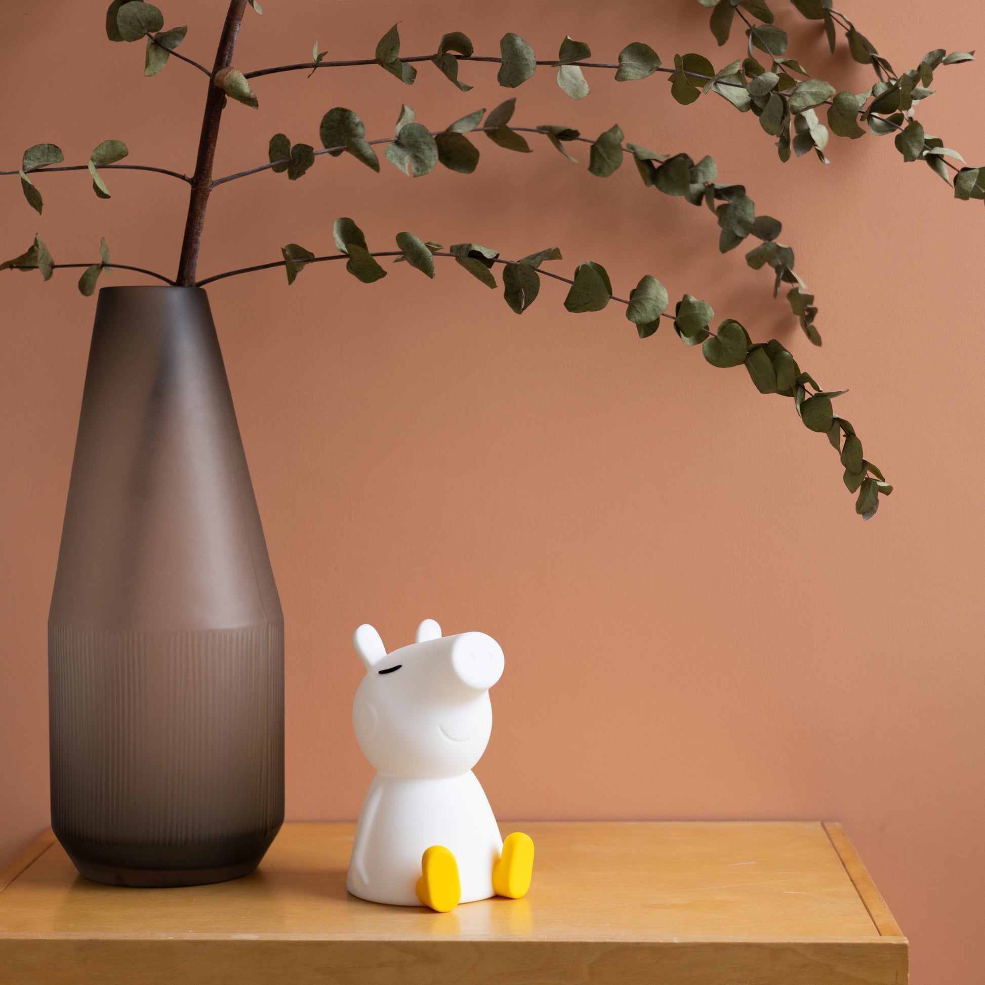 Decorative vase with a plant and a small white figurine on a wooden surface against a beige wall.