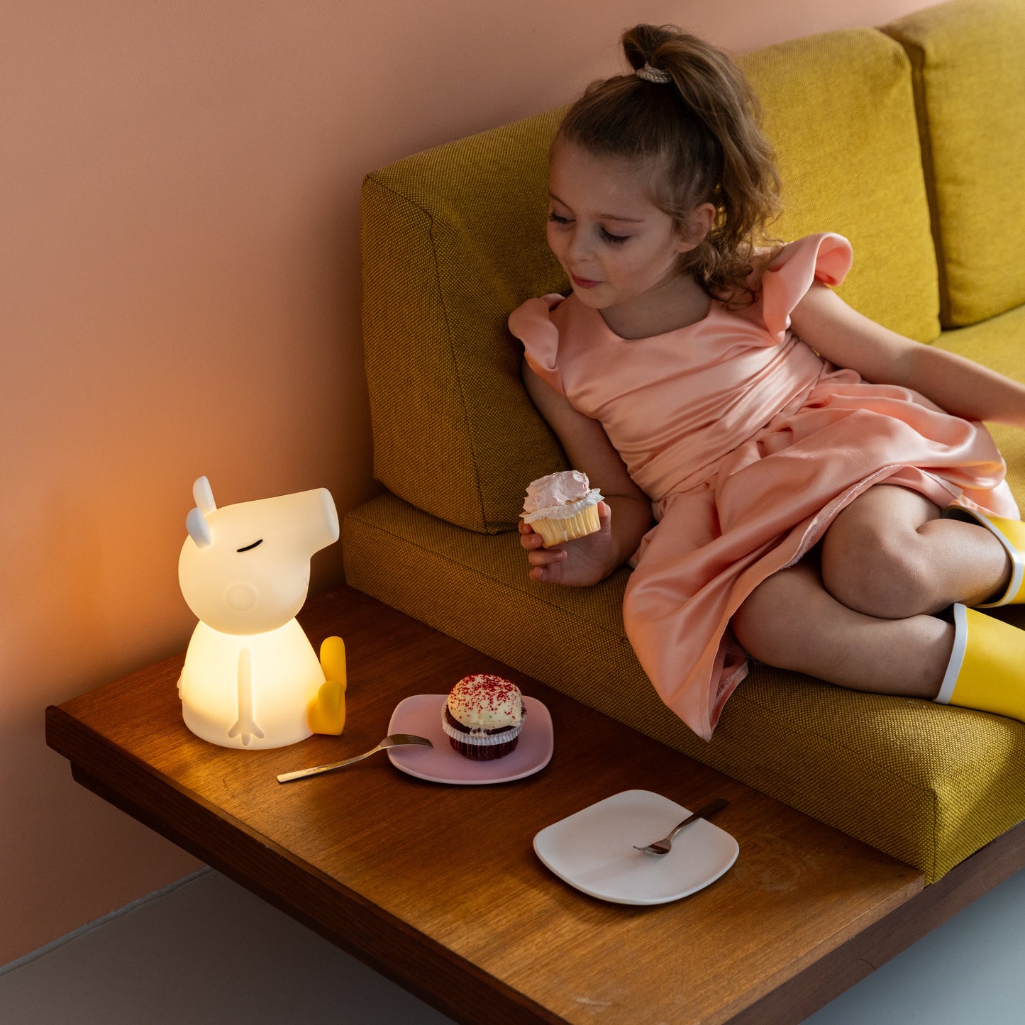 Young girl sitting on a yellow couch with a small table beside her, holding a cupcake.