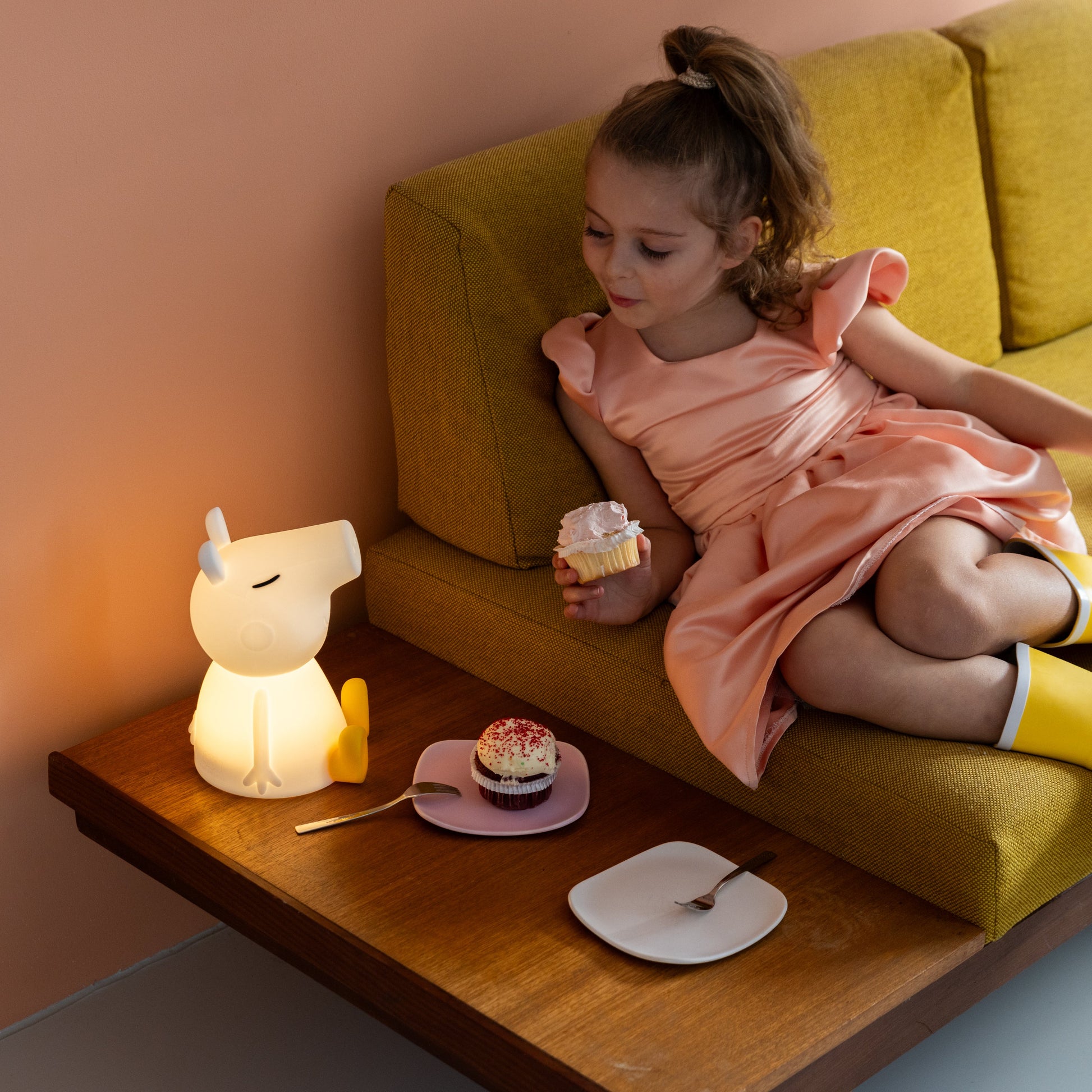Young girl sitting on a yellow couch with a small table beside her, holding a cupcake.