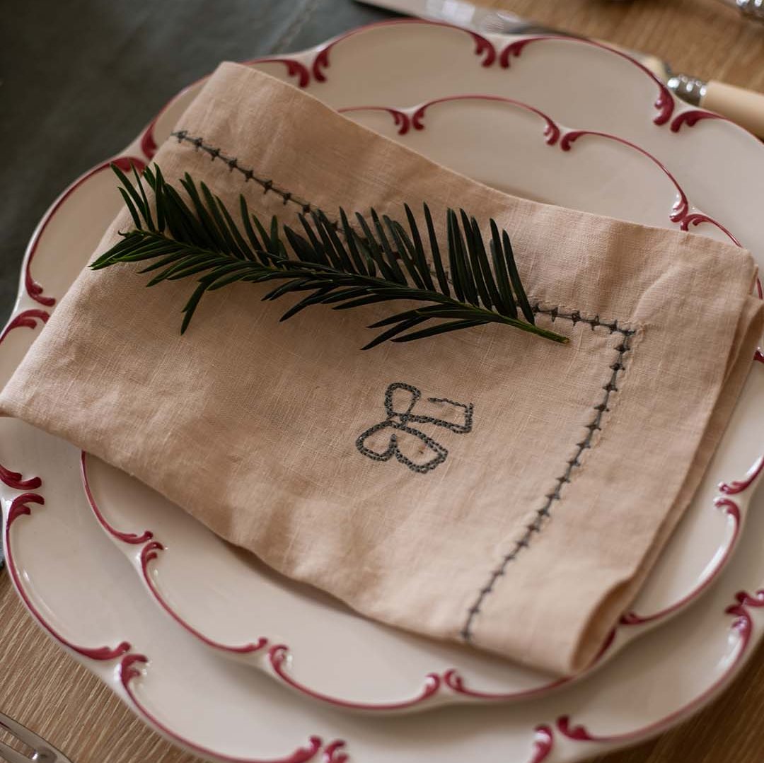 Folded beige napkin with embroidery on a set of white plates with red trim.