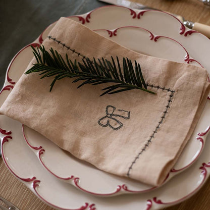 Folded beige napkin with embroidery on a set of white plates with red trim.