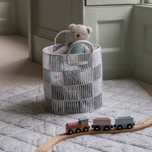 Woven basket with a teddy bear on a textured rug next to a wooden train set