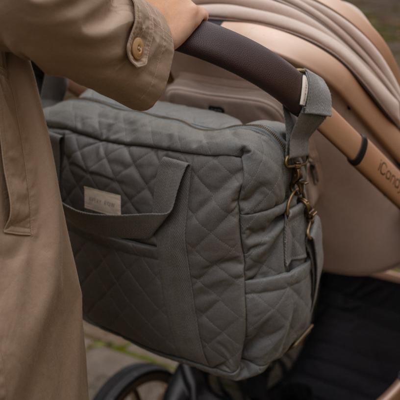 Gray quilted bag attached to a stroller with a person holding onto it.
