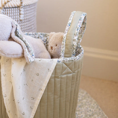Quilted baby basket with floral trim containing a teddy bear and blanket on a neutral background