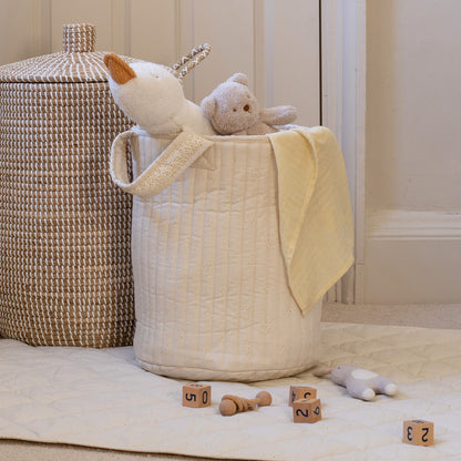 White textured laundry basket with toys and a blanket on a light wooden floor.