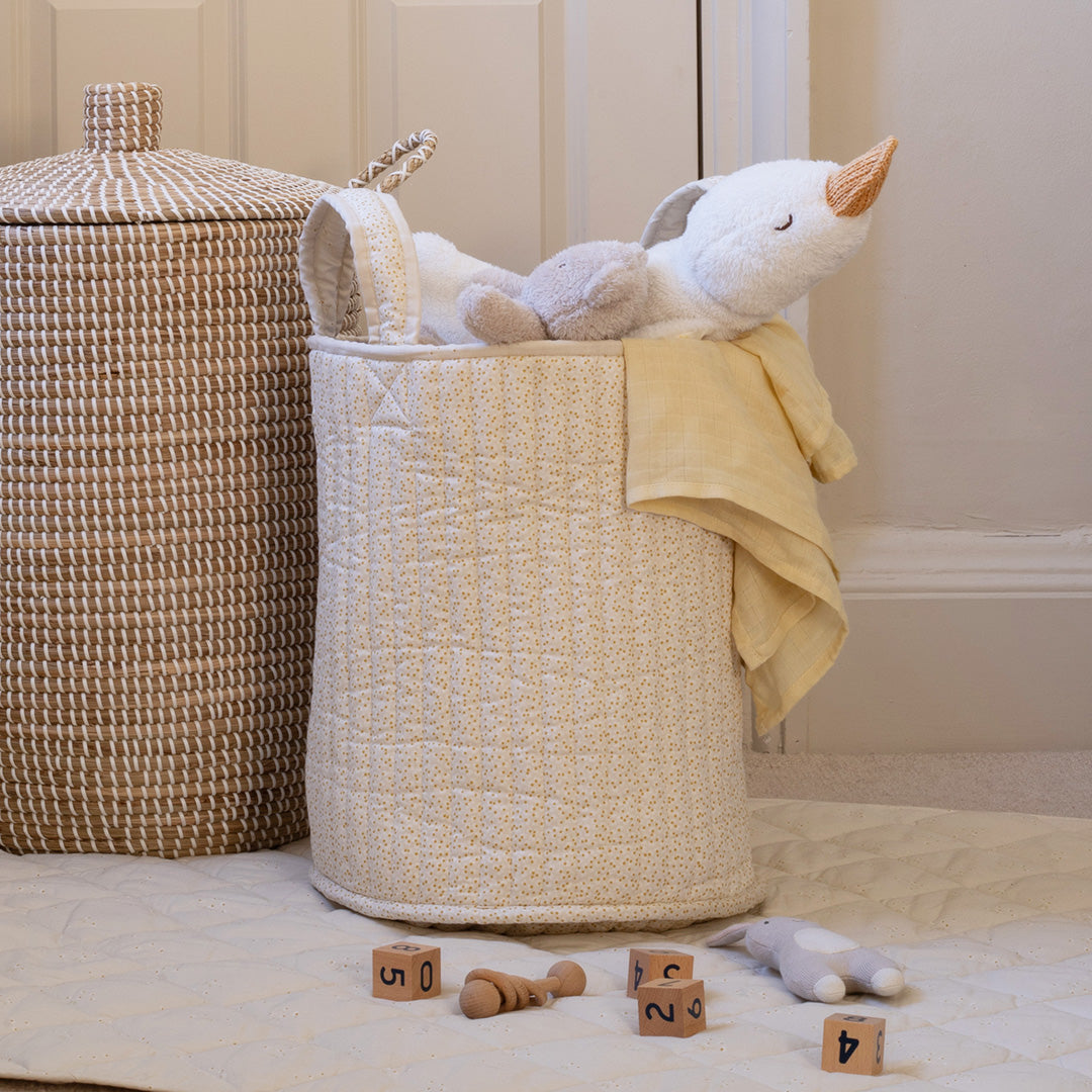 Beige woven laundry basket with toys and a towel on a wooden floor.