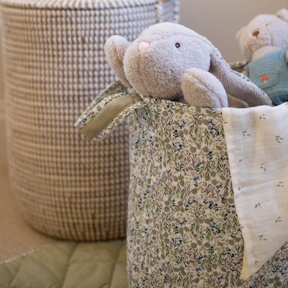 Floral-patterned basket with stuffed animals and a blanket on a bed.
