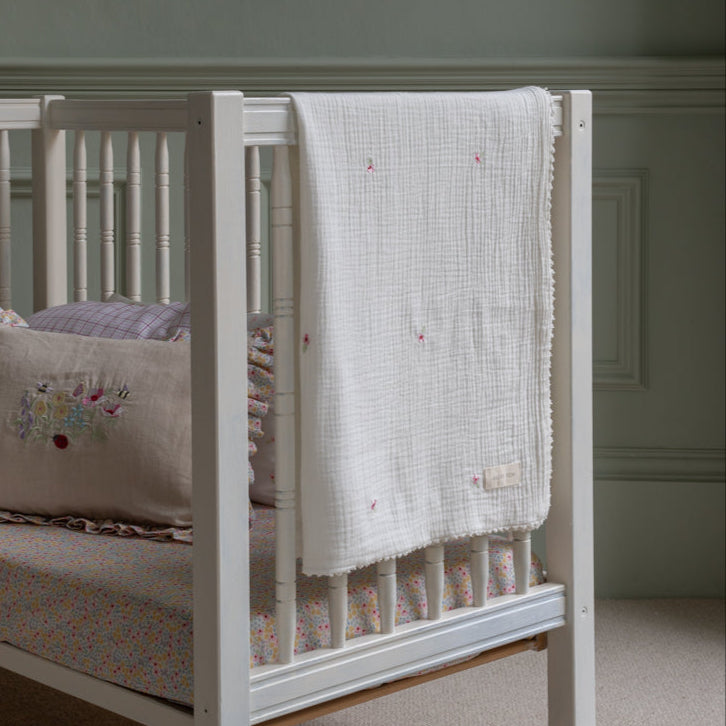 White crib with a floral mattress cover and embroidered blanket in a room.