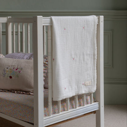 White crib with a floral mattress cover and embroidered blanket in a room.