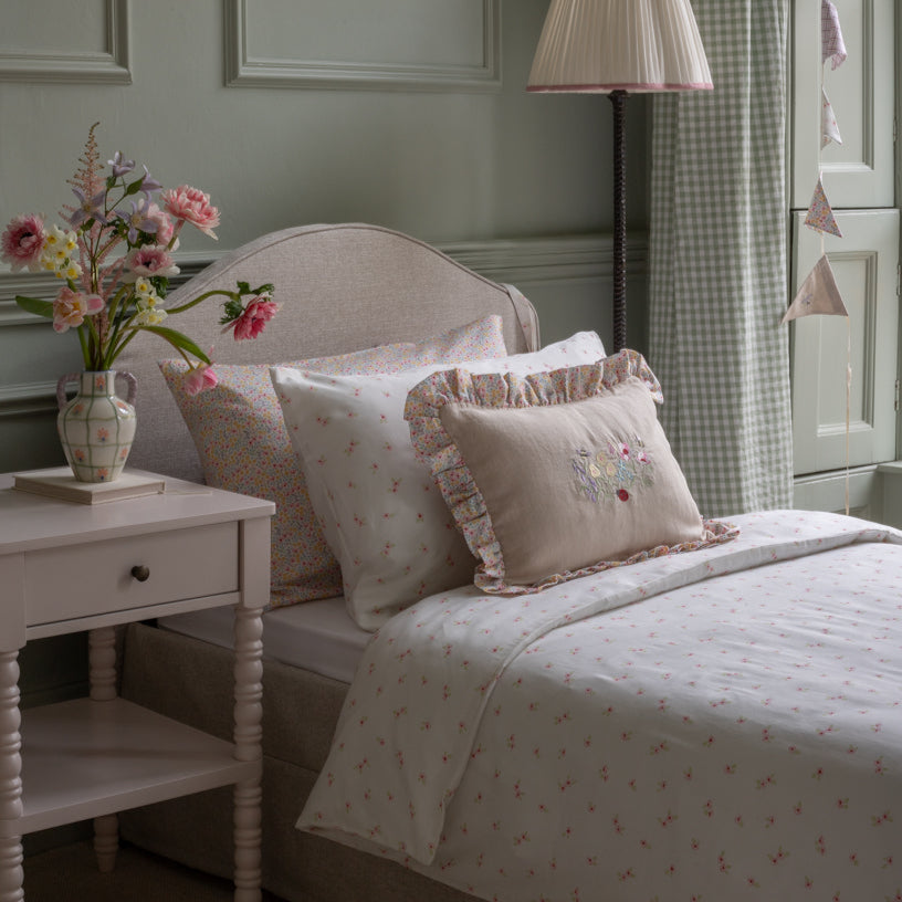 Neatly made bed with decorative pillows and a vase of flowers on a nightstand in a bedroom.