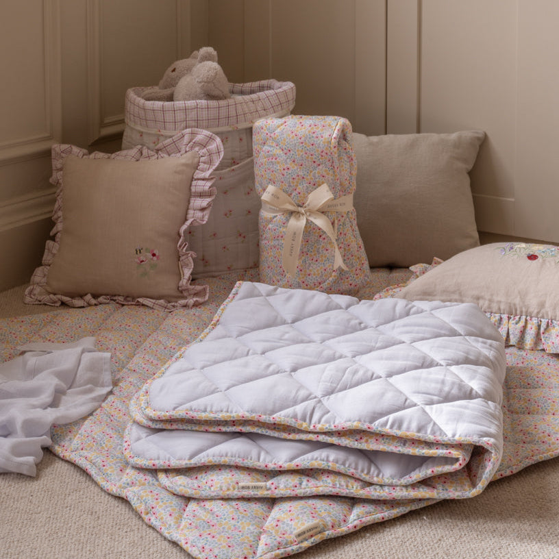 Stack of floral-patterned quilts on a bed with pillows and a teddy bear in the background.