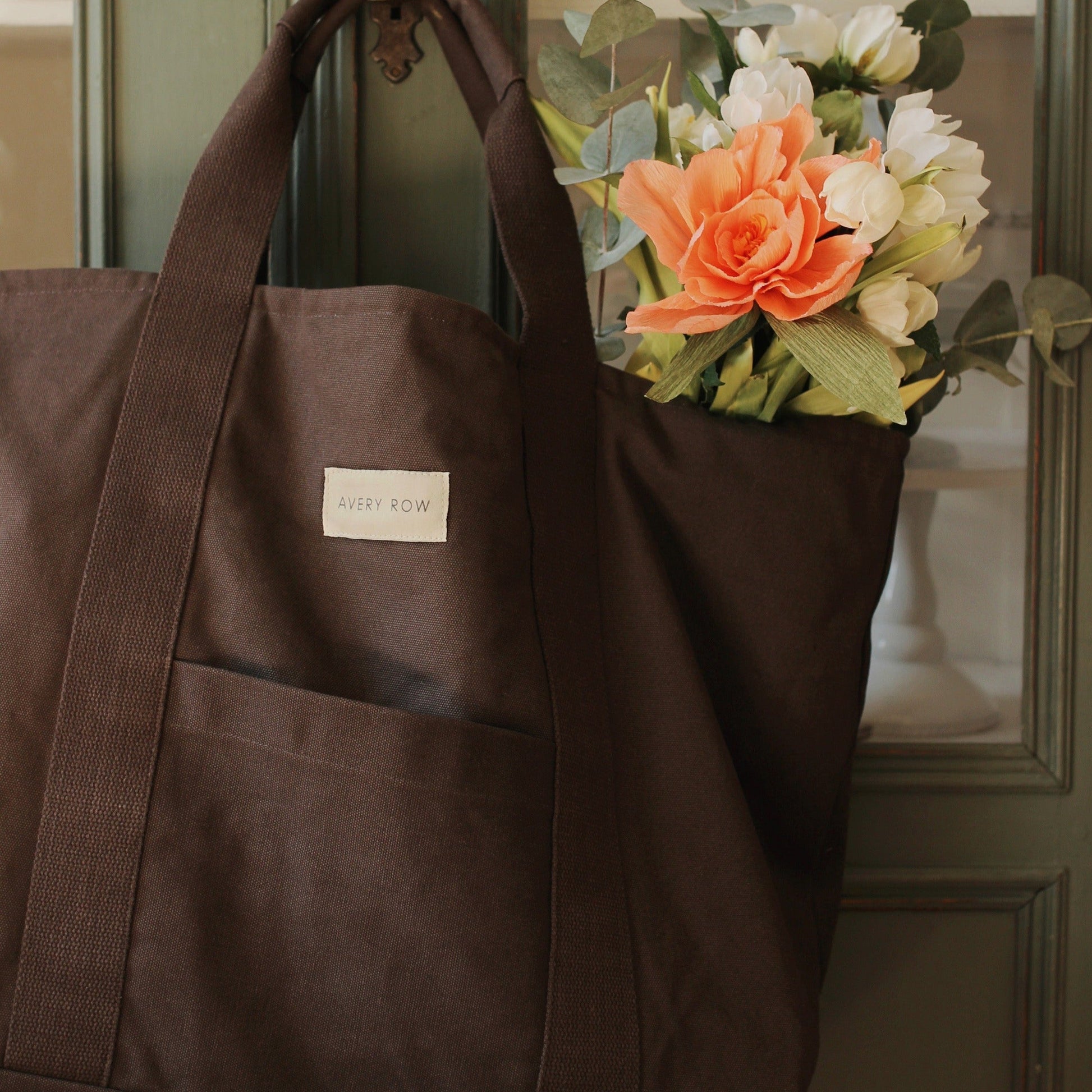 Brown tote bag with a visible brand label, held open to reveal a bouquet of flowers.