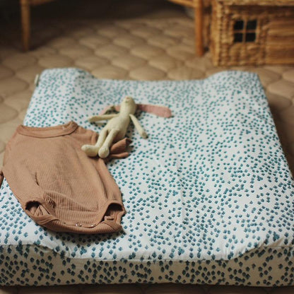 Brown baby romper and toy on a patterned changing pad with a wooden toy house in the background.