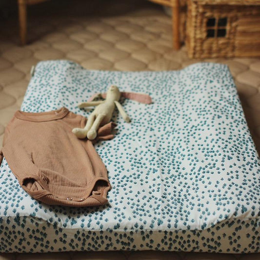 Brown baby romper and toy on a patterned changing pad with a wooden toy house in the background.