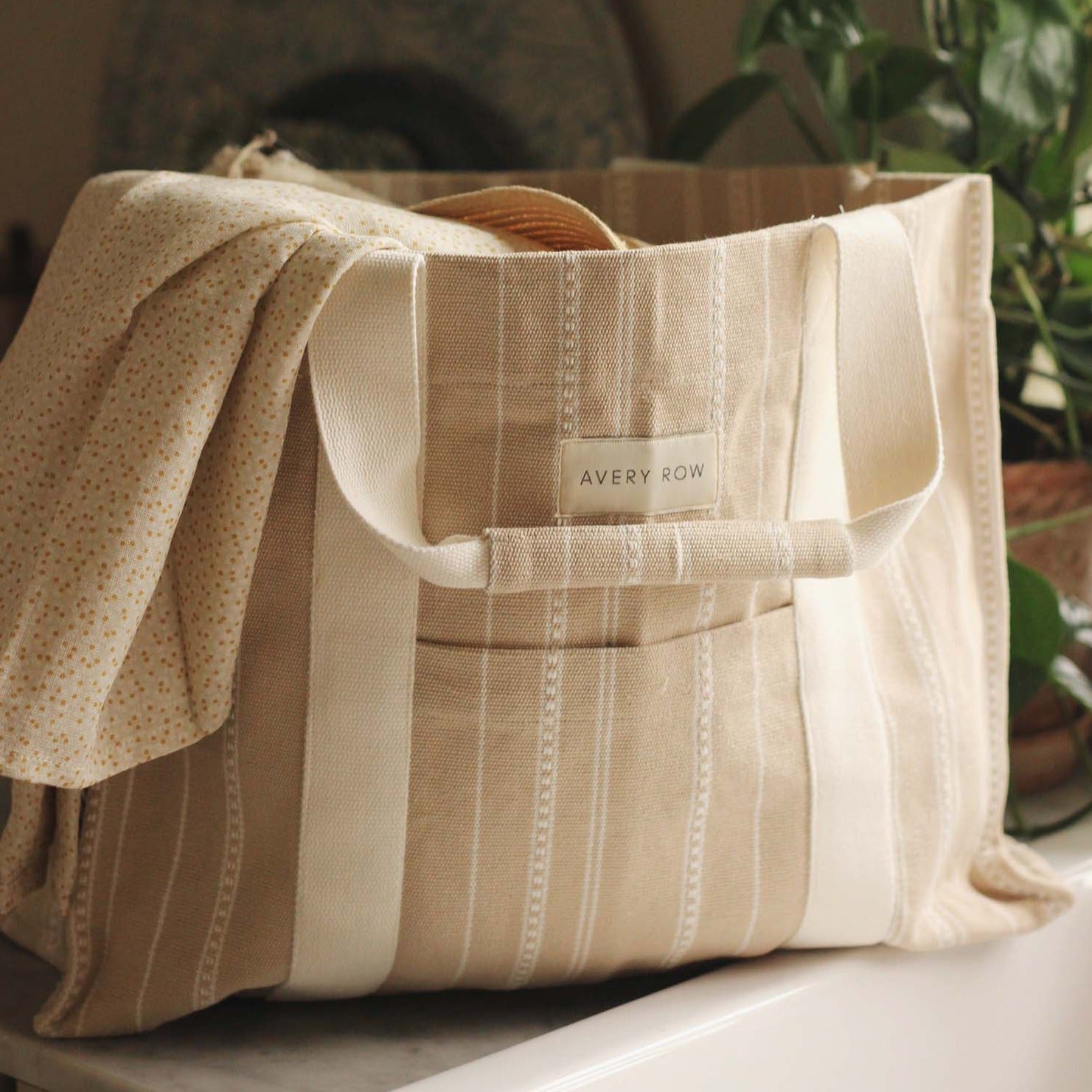 Beige striped tote bag with 'Avery Row' label on a surface with plants in the background