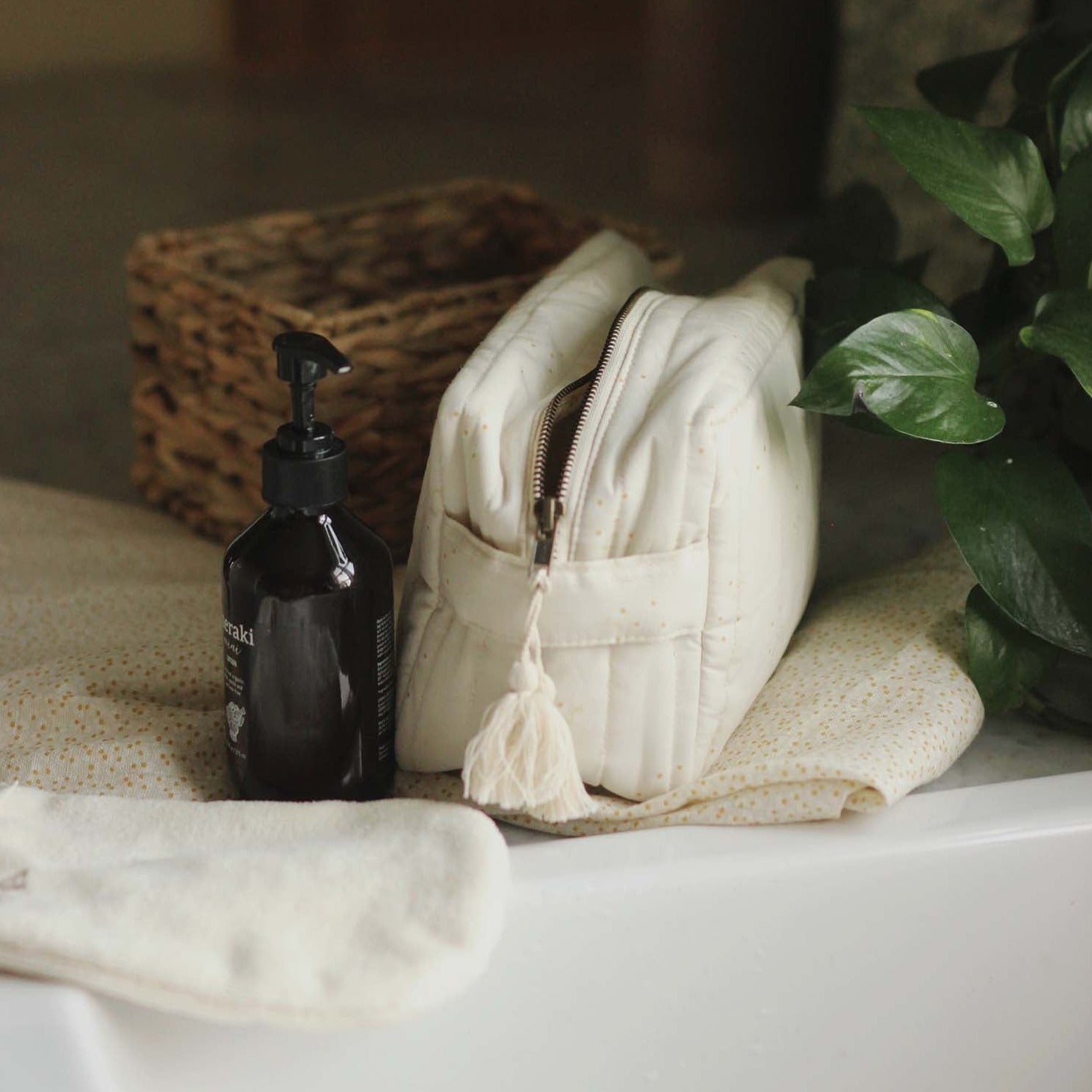 Black bottle with pump, white zippered pouch, and white towel on a surface with a plant in the background