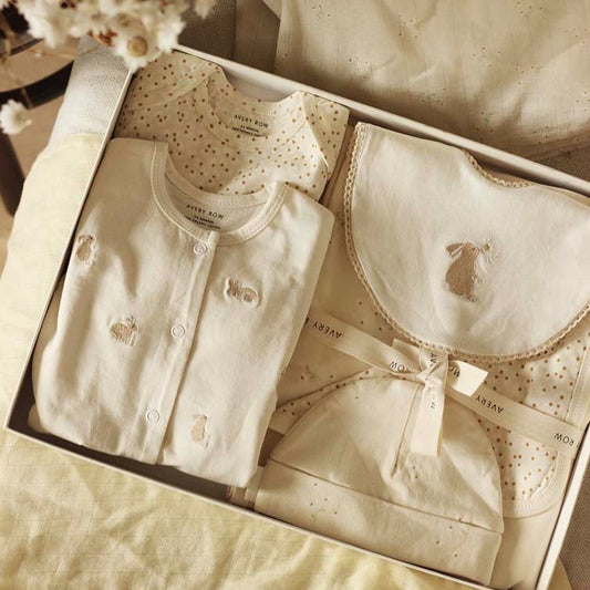 Baby clothing set with shirt, bib, and hat in a box on a soft beige background