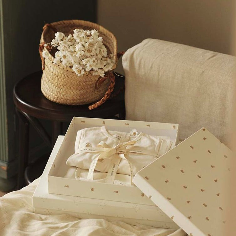 Baby clothes in a box on a bed with a basket of flowers in the background