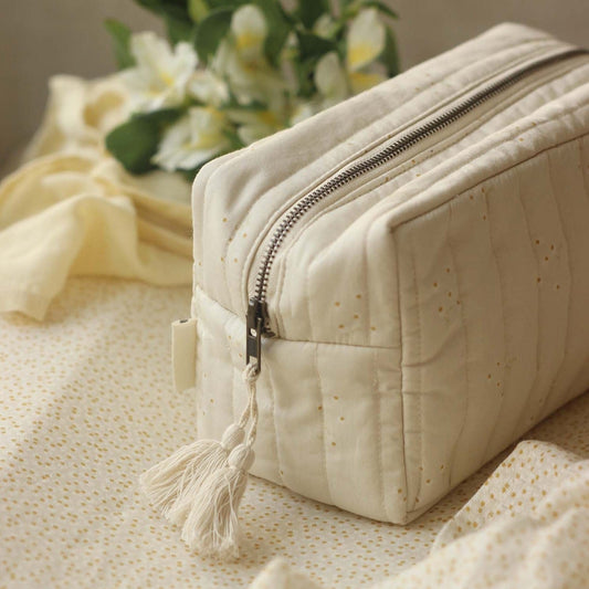 Beige toiletry bag with a tassel on a textured surface with flowers in the background