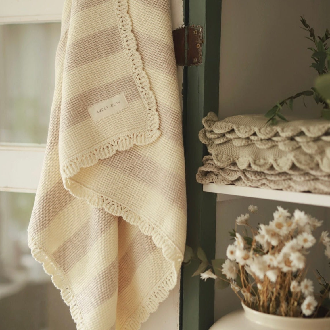 Knitted blanket with a brand label draped over a surface next to folded blankets and a vase with flowers.