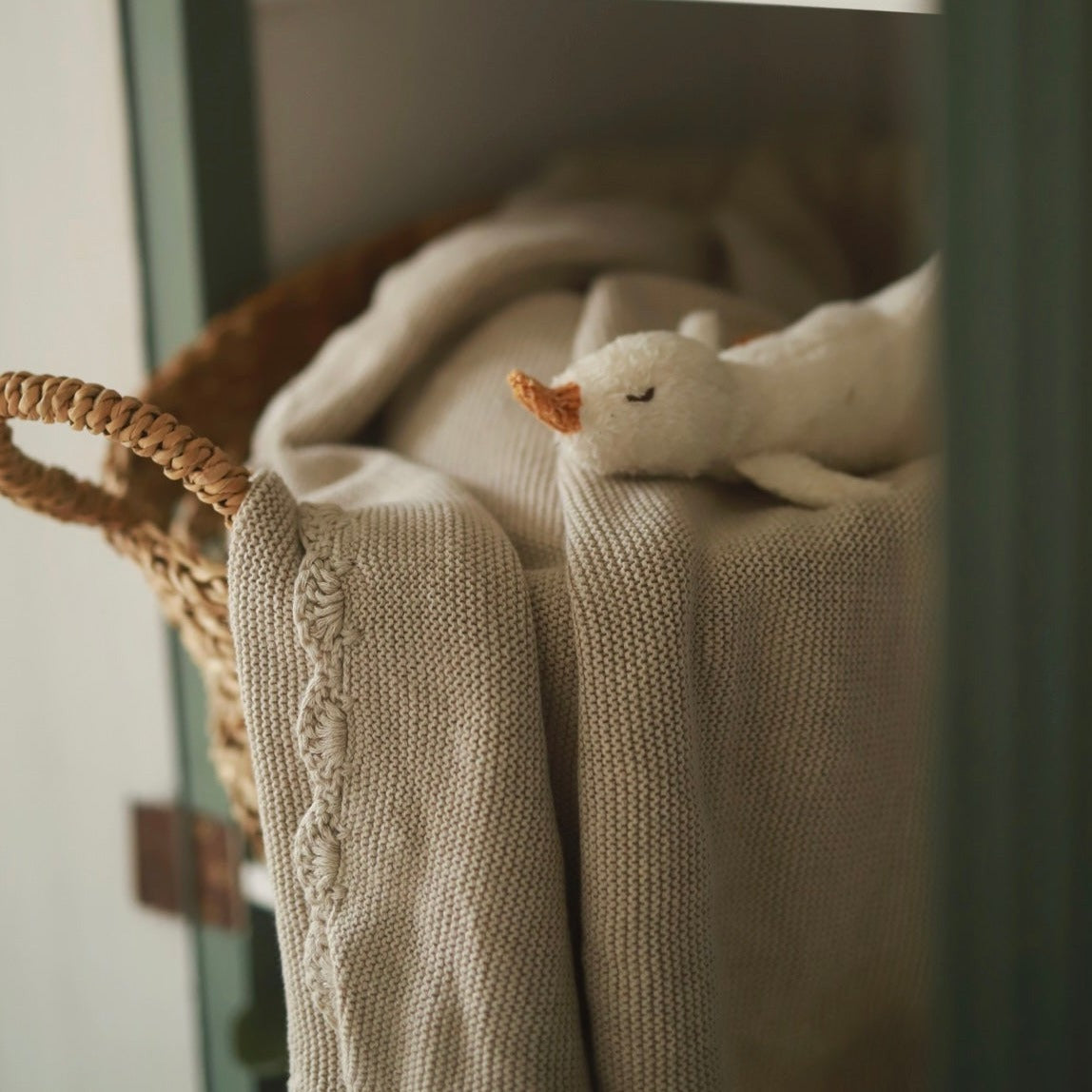 Close-up of a beige knitted blanket with a small white duck toy on top, next to a woven basket.