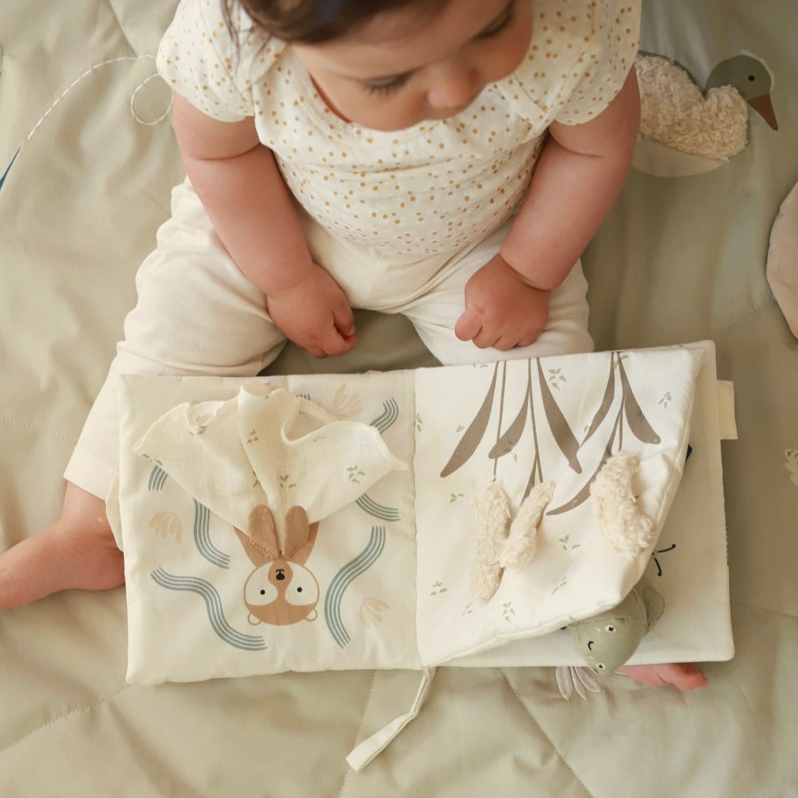 Baby sitting on a soft surface with a fabric book featuring animal illustrations.