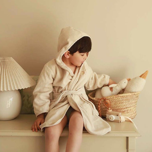 Child in a beige robe sitting on a table with toys, wearing a hooded robe.