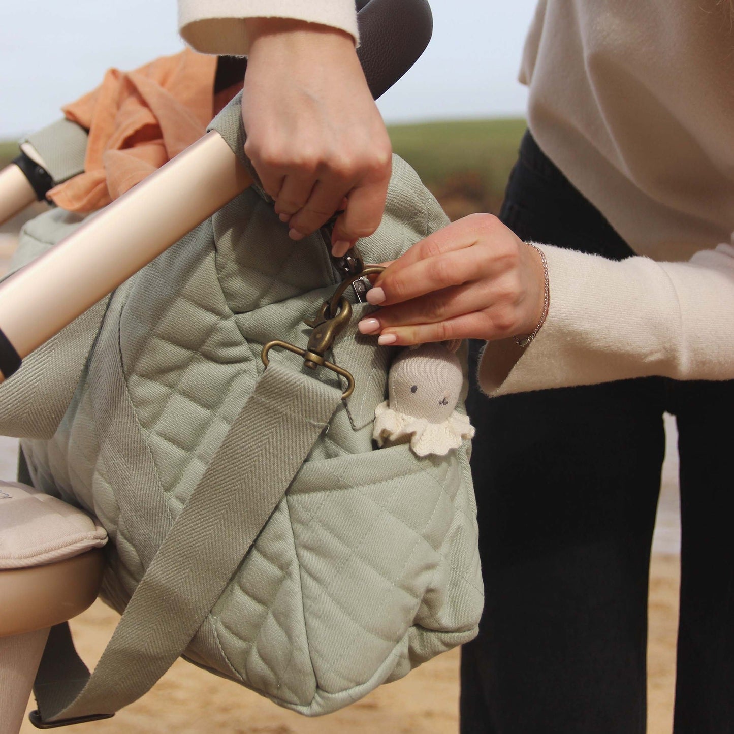 Person holding a green quilted bag with a small plush toy attached, outdoors.