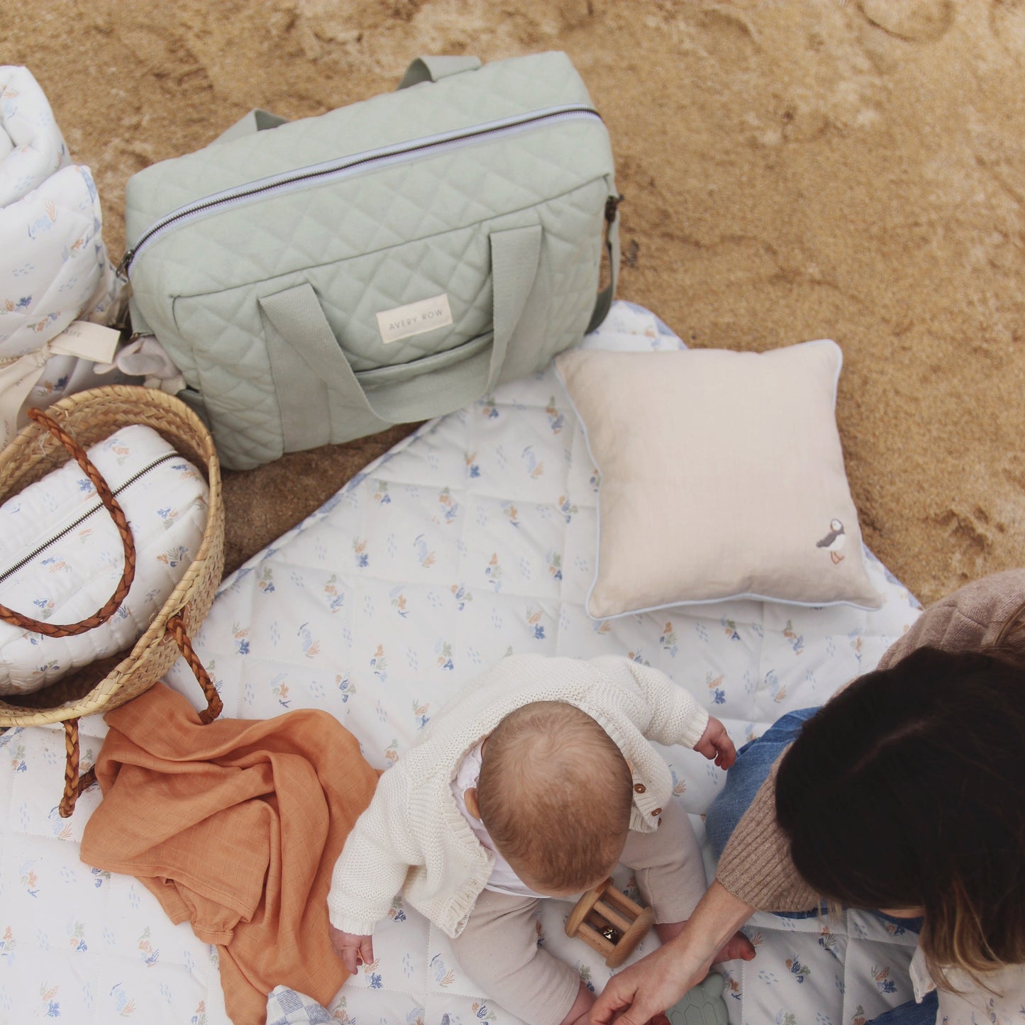 Baby and adult on a blanket with bags and pillows on sand