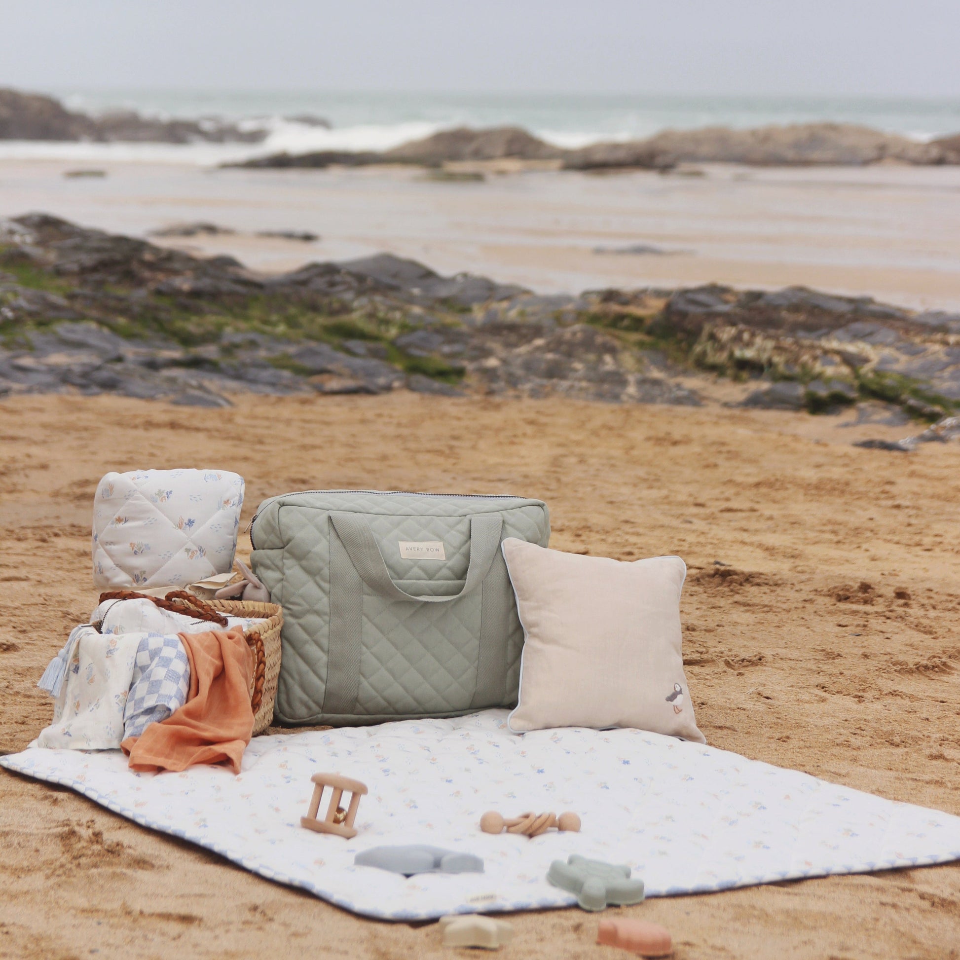 Outdoor setting with a quilted bag, pillows, and toys on a blanket by a rocky beach.